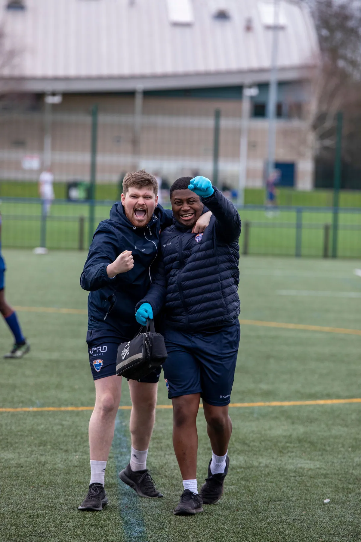 Two young men in team jackets celebrating together on a football pitch in Manchester